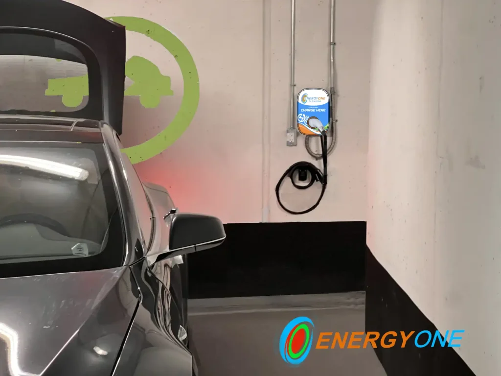 Electric vehicle parked in a charging station with the trunk open. A charging point labeled Energy One is mounted on the wall with cables hanging.