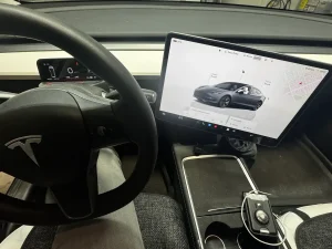 Interior view of a Tesla car showing the steering wheel and a touchscreen display with vehicle controls and navigation.