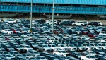 A large parking lot filled with rows of parked cars, predominantly white, under a blue-tinted glass structure.