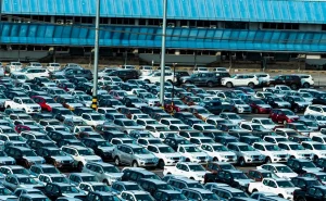 A large parking lot filled with rows of parked cars, predominantly white, under a blue-tinted glass structure.